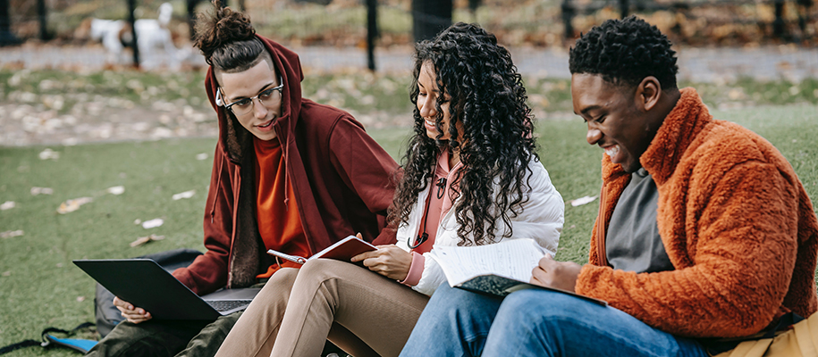 Three students in fall jackets sitting in grass with notebooks, bags, and laptop