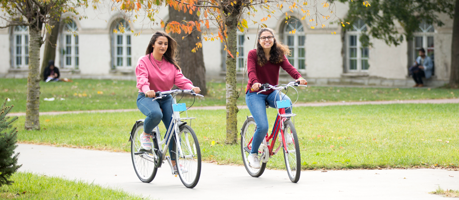 Two White females smiling and riding bicycles on college campus in the fall