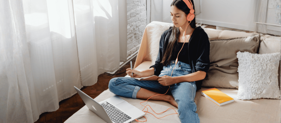 Young woman sits on a couch with a laptop, writing while wearing headphones