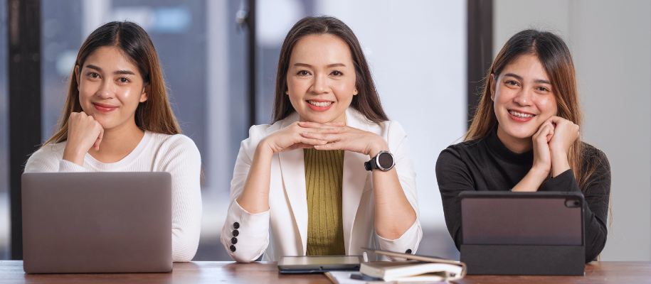 Asian woman in white suit smiling between two younger Asian women with tablets