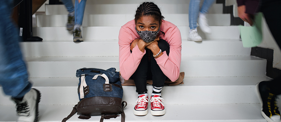 Black woman with face mask hugging knees next to backpack on busy school stairs