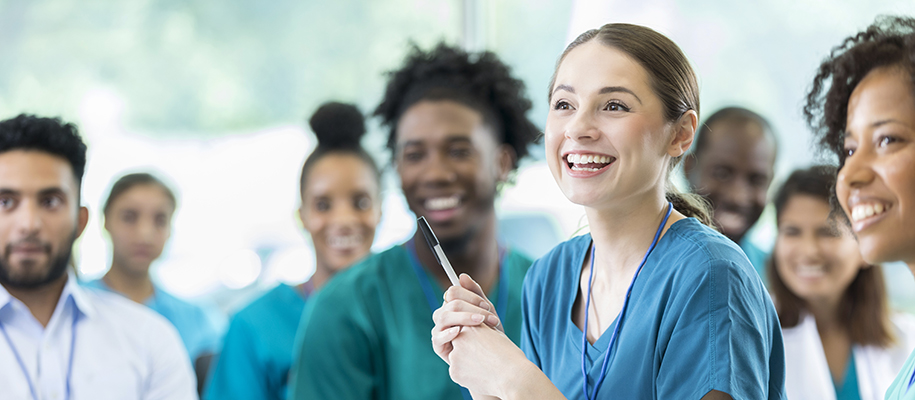 White woman in blue scrubs holding pen, smiling in class full of medical student