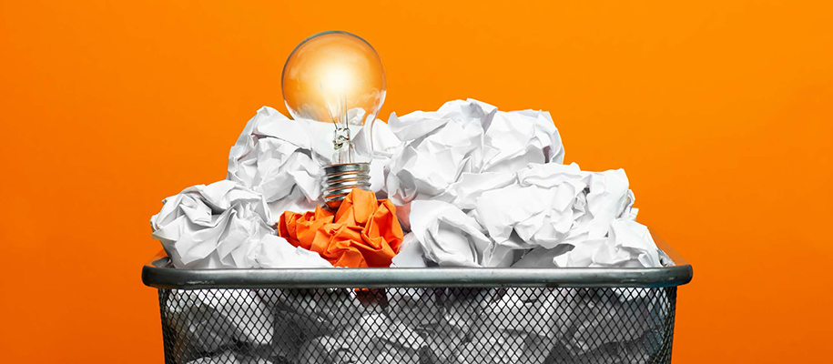 Lit lightbulb in trash can of crumpled white paper, one crumpled orange page