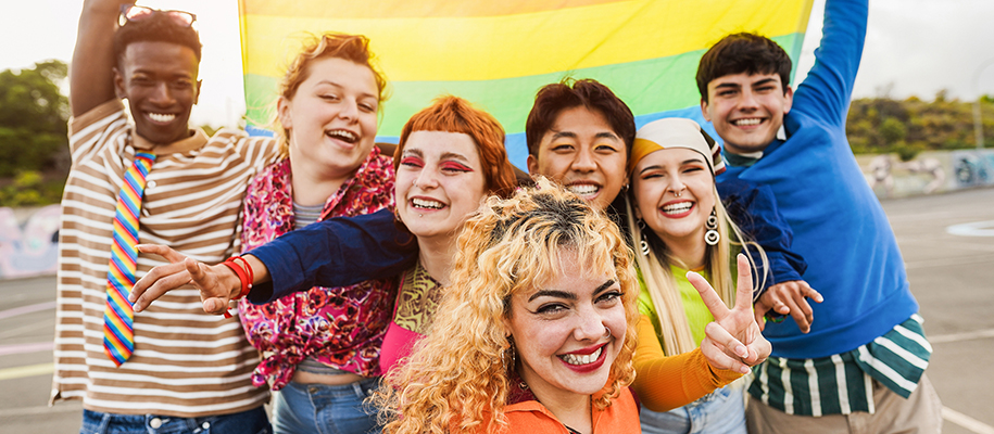 Group of queer friends in bright clothing, smiling and holding up pride flag