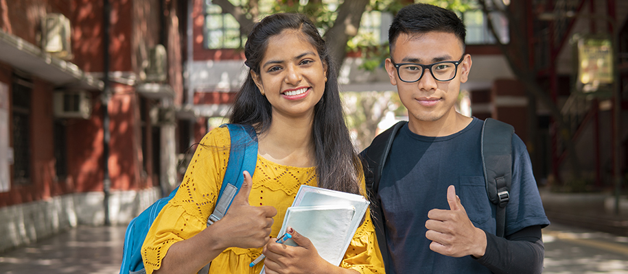 South Asian woman in yellow, east Asian man in glasses, outside giving thumbs up
