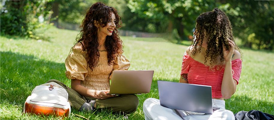 Two Latina women with curly hair in park with laptops doing online work together