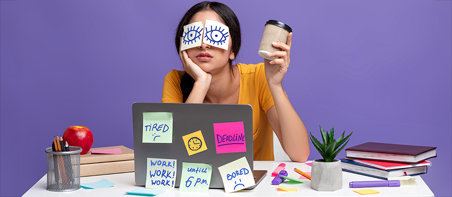 Indian woman with sticky note eyes over face, holding paper cup at messy desk