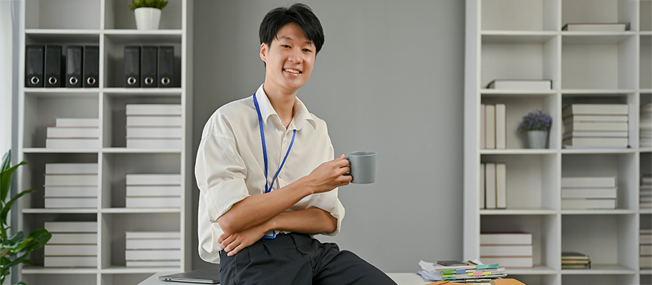 Asian man in white shirt, smiling and holding coffee mug sitting on office desk