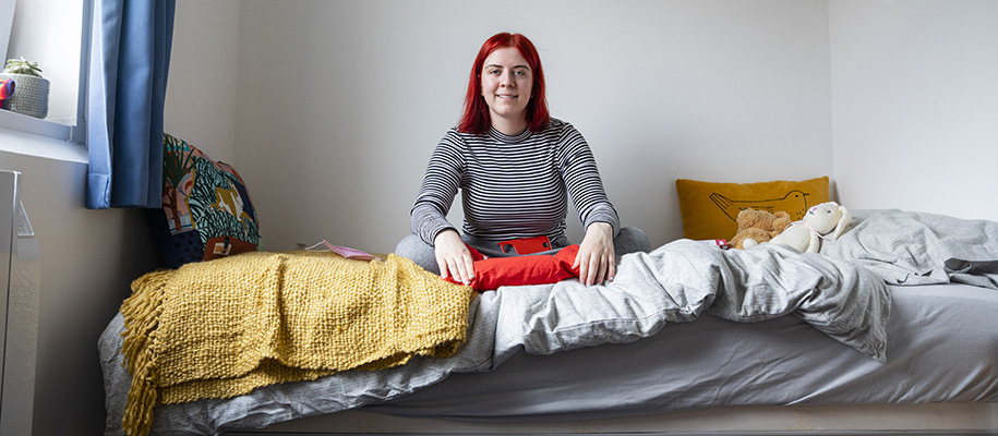 White woman with red hair in striped shirt sits on bed in college dorm room