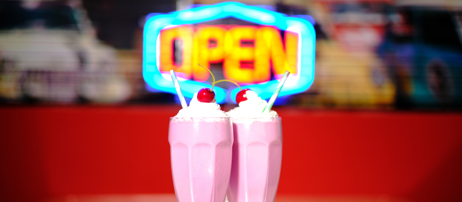 Two pink milkshakes with whipped cream & cherries in front of neon open sign