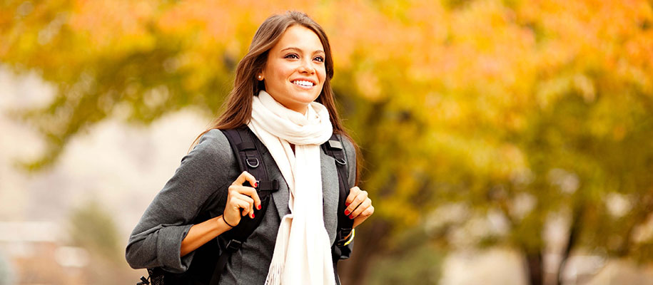 Brunette with scarf and backpack looking excited on campus with fall foliage