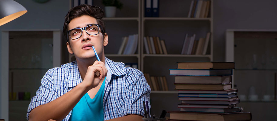 Dark-haired male wearing glasses, holding pencil to chin, thinking in study