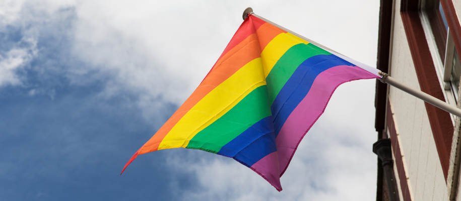 Rainbow Pride flag on pole attached to building against blue sky with clouds