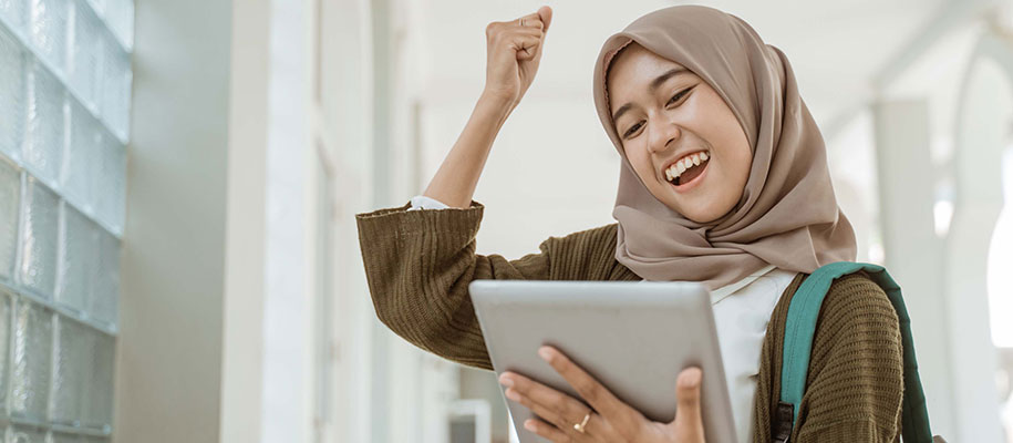 Happy Muslim female student holding tablet and raising fist in air excitedly