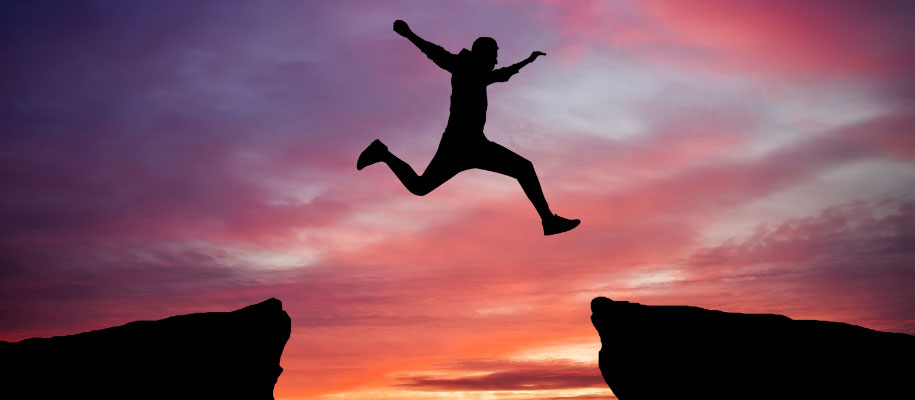 Silhouette of man jumping over gap from one cliff to another at sunset