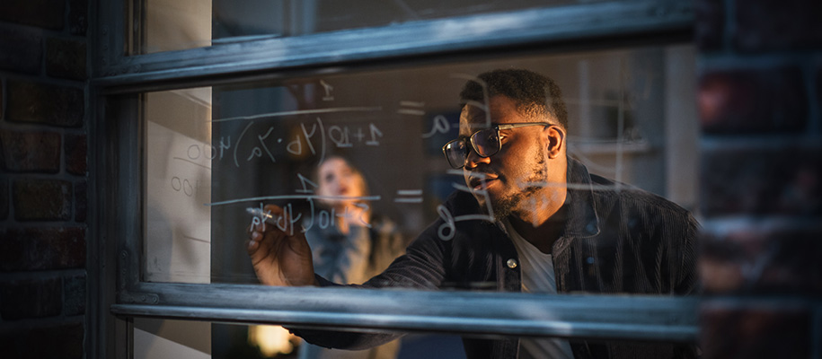 Black man in glasses writing equations on window, woman looking pensive behind