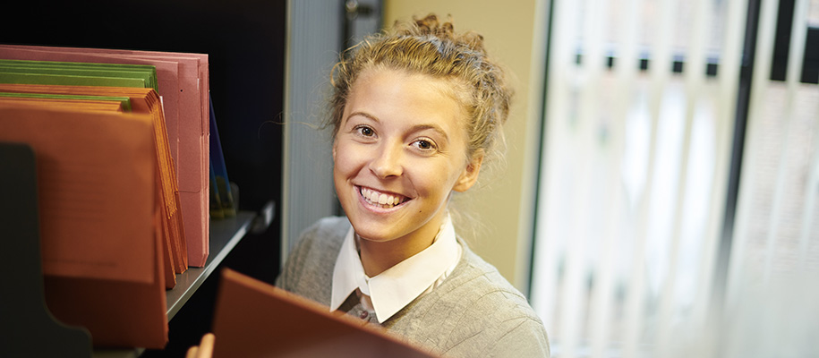 White woman in collared shirt smiling in office, holding file next to shelf