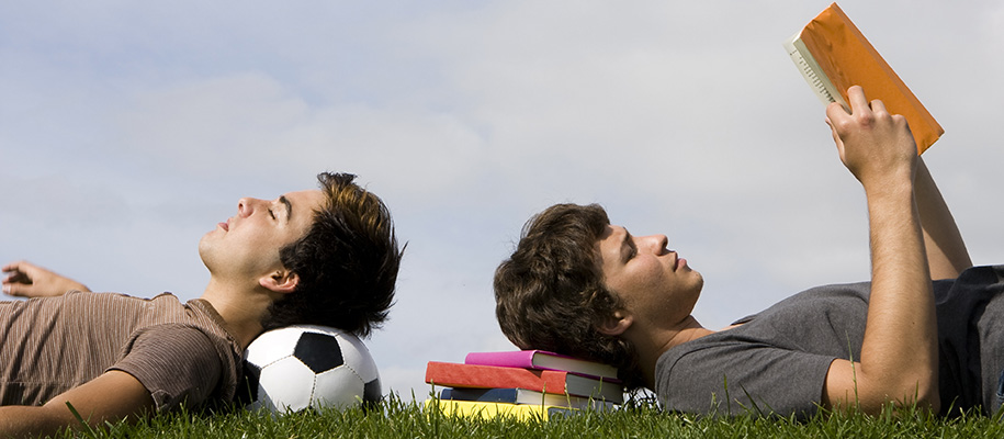 Two White teen boys, one laying on soccer ball, one on stack of books outside