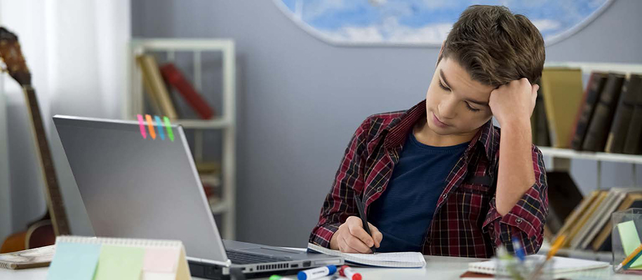 White male student in plaid shirt at home desk writing in notebook, head in hand