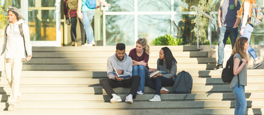 Three diverse students sitting on building steps with books while others walk by