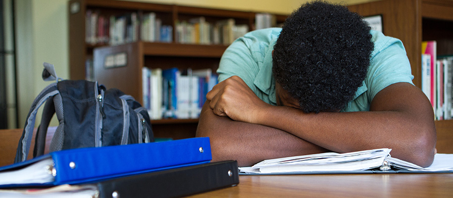 Black male teen with head down on arms in library with binders and backpack