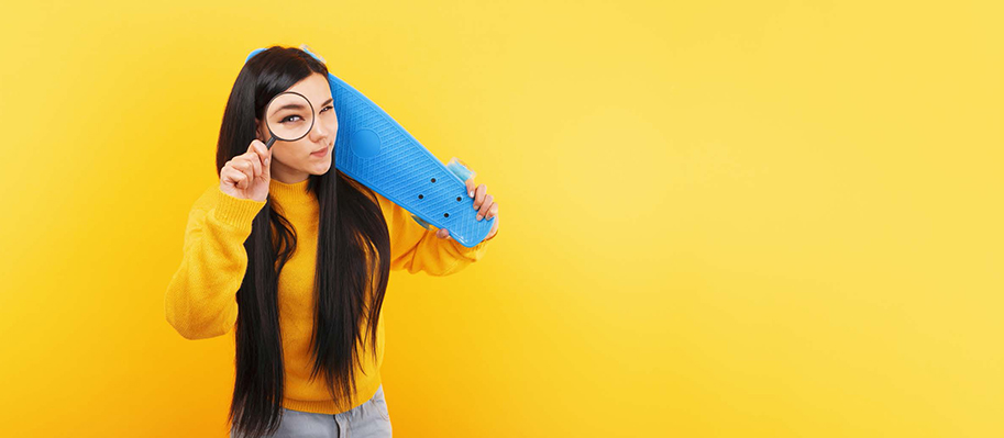 Asian woman in yellow sweater, with magnifying glass to eye and penny board