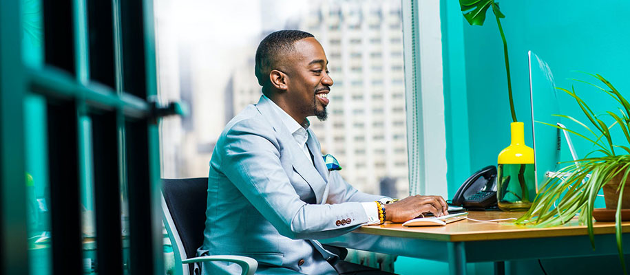 Black man in suit smiling at computer at desk next to window overlooking city