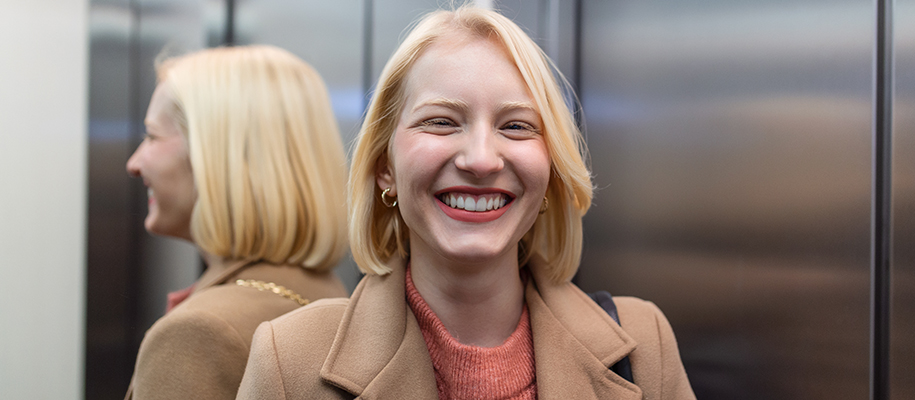 Blonde White woman in brown peacoat, smiling in elevator, reflection behind