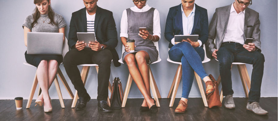 Five diverse businesspeople in line of chairs looking at phones, tablets, laptop