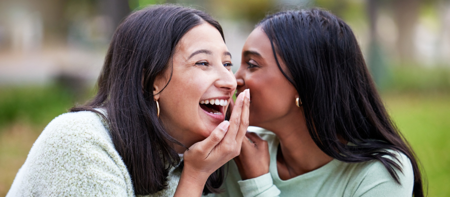 Two multicultural women laughing and whispering secrets on campus