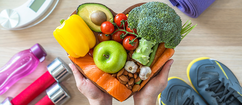 Hands holding heart of fruits, veggies, healthy proteins over workout equipment