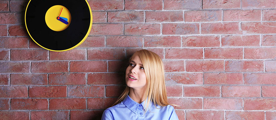 Blonde woman standing against brick wall looking up at black and yellow clock