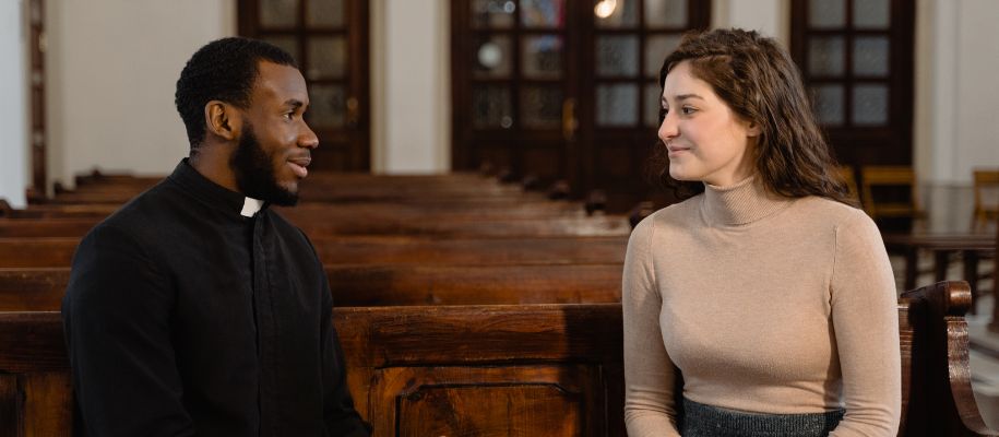 Young Black male priest sitting in pew with young White woman in turtleneck