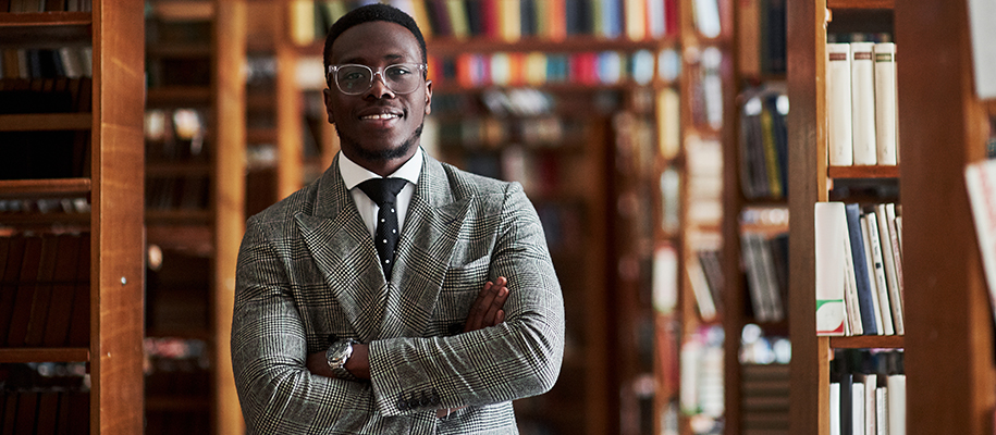 Black man in glasses, business suit, watch in library with packed shelves