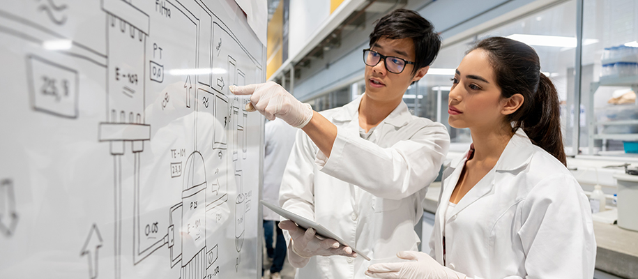 Asian man and Hispanic woman in lab coats focused on white board diagram