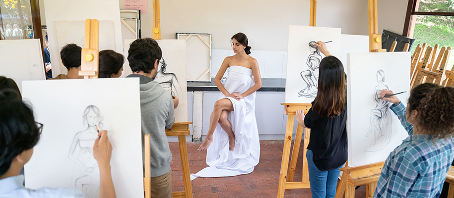 Art class standing at easels drawing live model in draped white dress