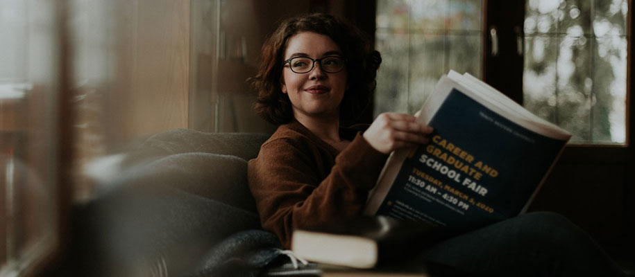 Short-haired brunette in glasses sitting with grad school fair book looking away