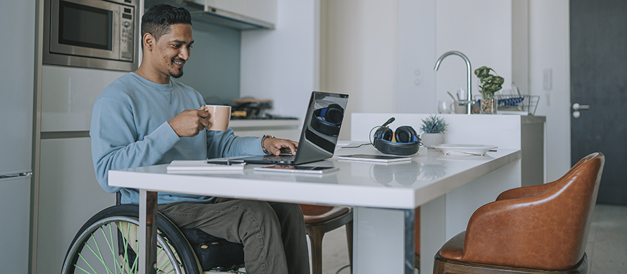 Hispanic man wheelchair in kitchen, drinking coffee and working on computer