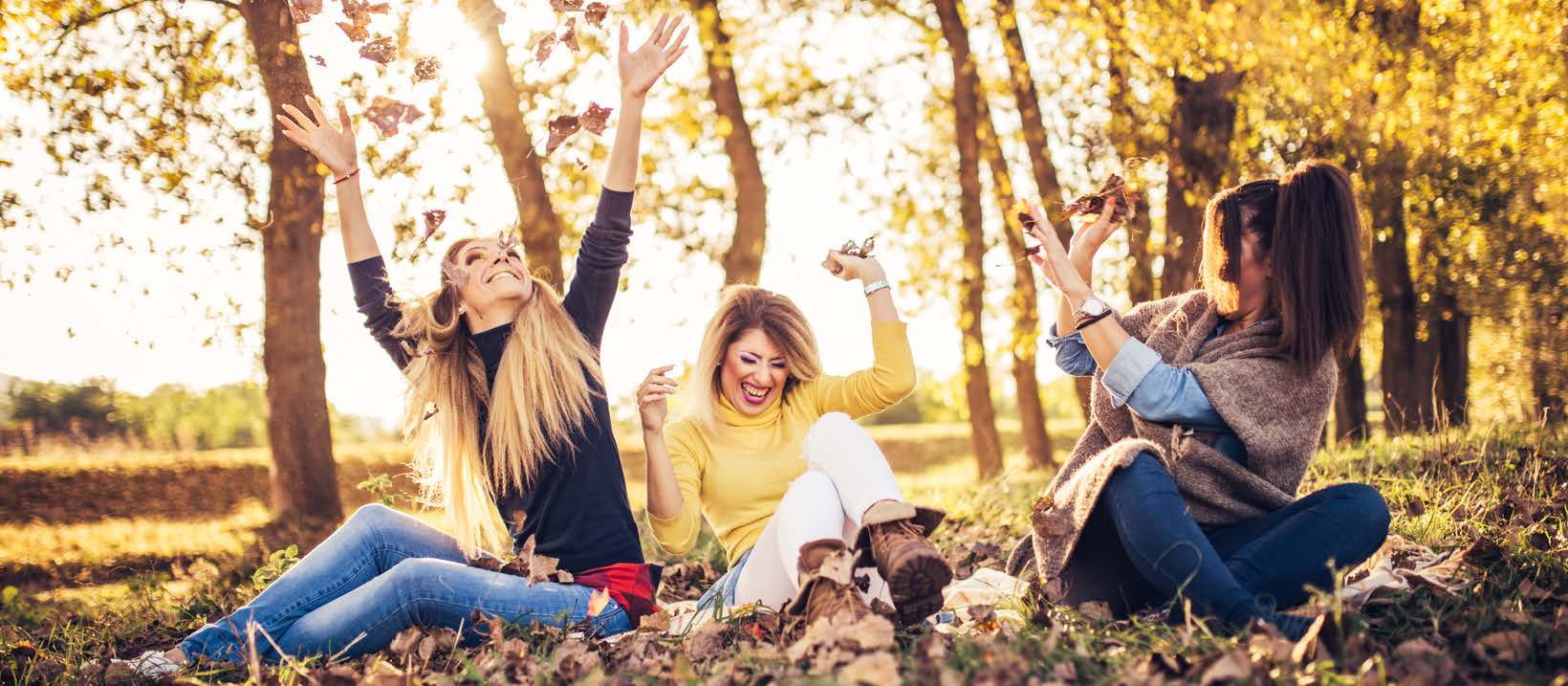 Three White women sitting in park throwing leaves in air and laughing