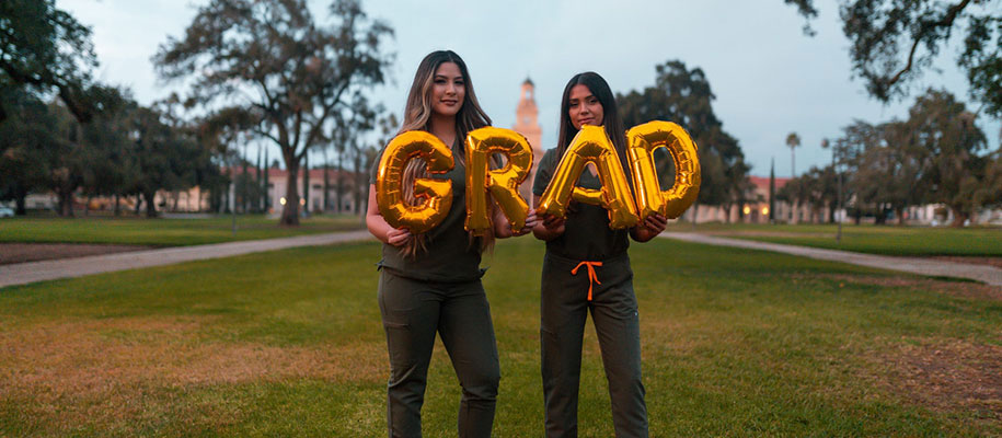 Two dark-haired females standing on campus lawn holding gold Grad letter balloon