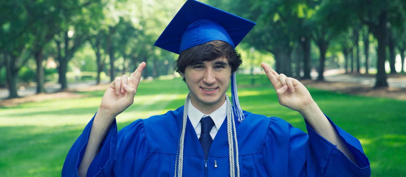 White man in blue grad cap and gown outside crossing fingers and smiling