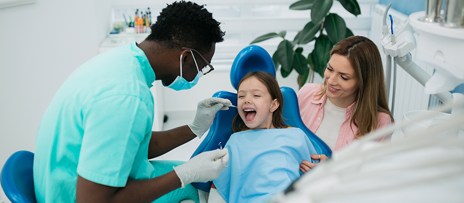 Young Black dental hygienist using dental tools on young girl, mom beside