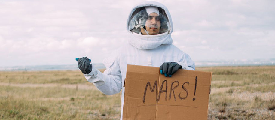 White male in astronaut suit hitchhiking with cardboard sign reading Mars