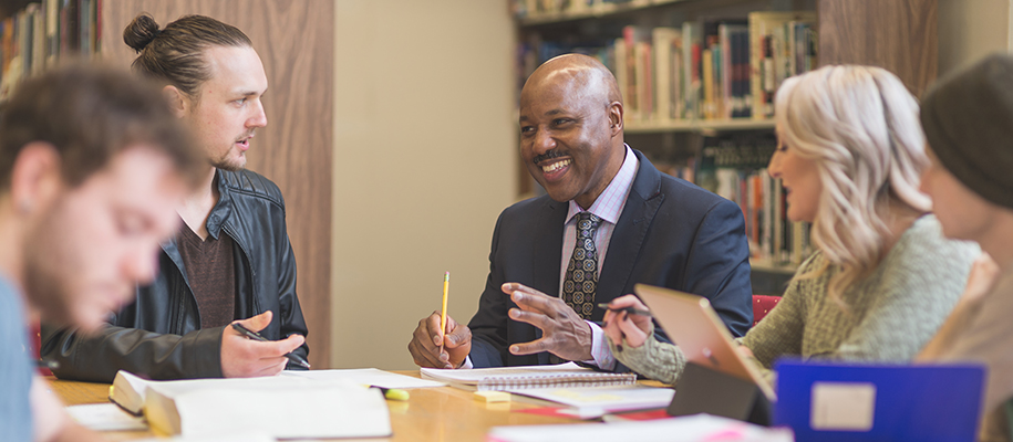 Black male professor in suit collaborating with group of students in library