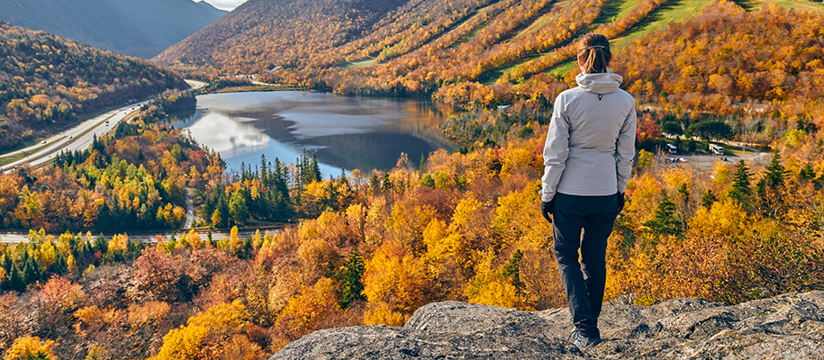 Woman in grey winter coat standing on rock overlooking fall trees on mountains