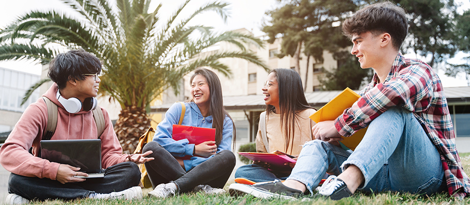 Four diverse international students sitting on campus lawn with books, backpacks