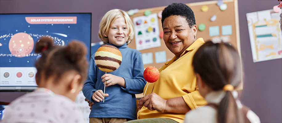 Happy Black woman, blond boy hold planets on sticks in class with other kids