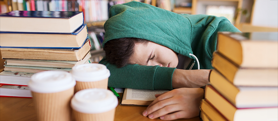 White male student in green sweatshirt asleep surrounded by books and coffee