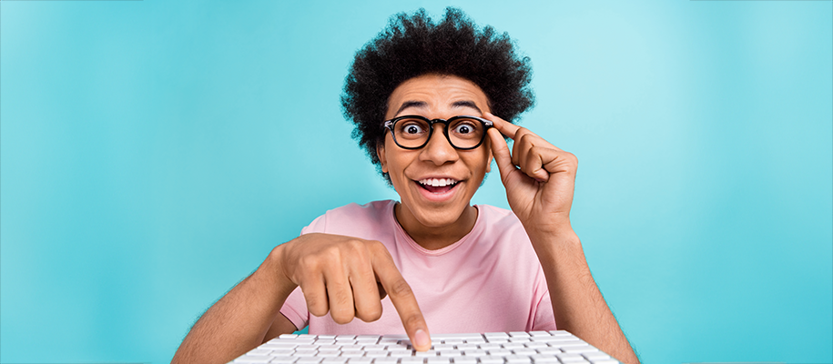 Black male teen with black glasses presses button on keyboard on blue backdrop