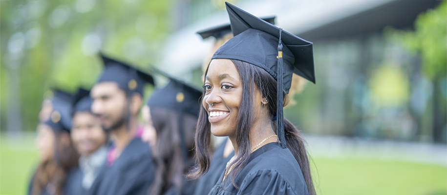 Black woman with nose ring in black cap and gown, smiling at end of row of grads